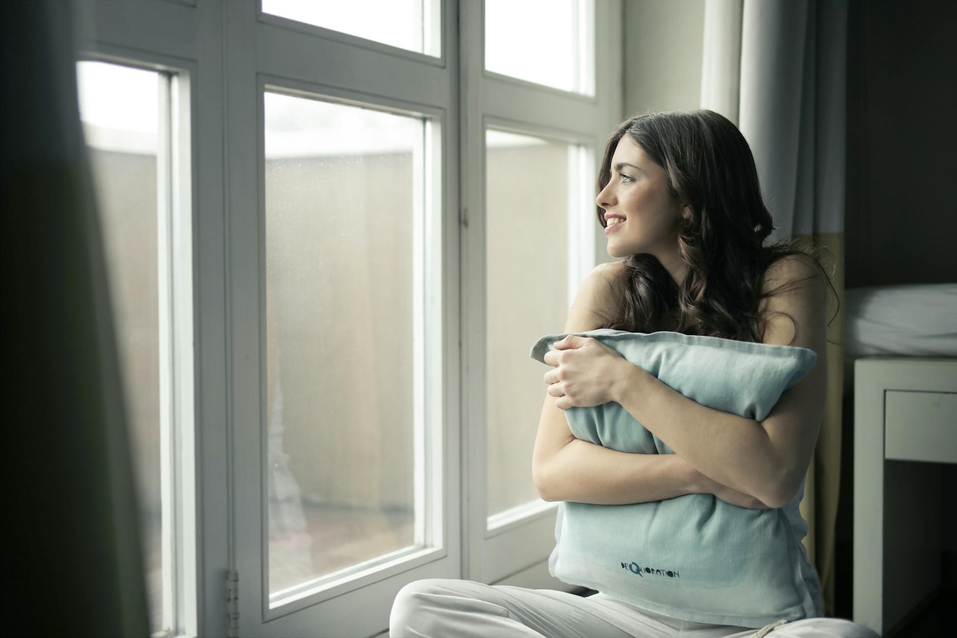 black haired woman hugging gray pillow near glass panel window